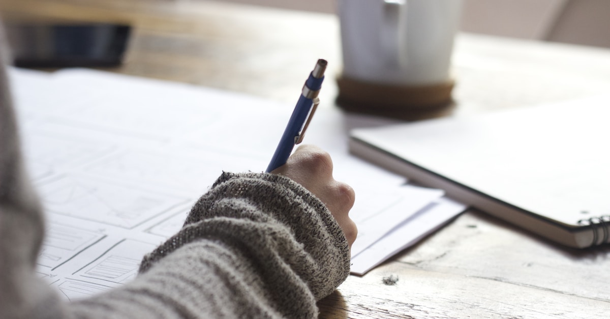 Person writing on brown wooden table near white ceramic mug
